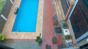 Ein rechteckiger Swimmingpool mit Holzterrasse, umgeben von einem roten Ziegelpfad und Blumentöpfen.