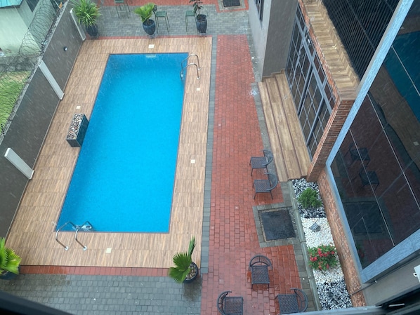 A rectangular swimming pool with a wooden deck, surrounded by a red brick pathway and potted plants. - Meridian Residence (Tema)