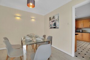 A dining area with a glass table set for six, surrounded by chairs, with a kitchen visible in the background.