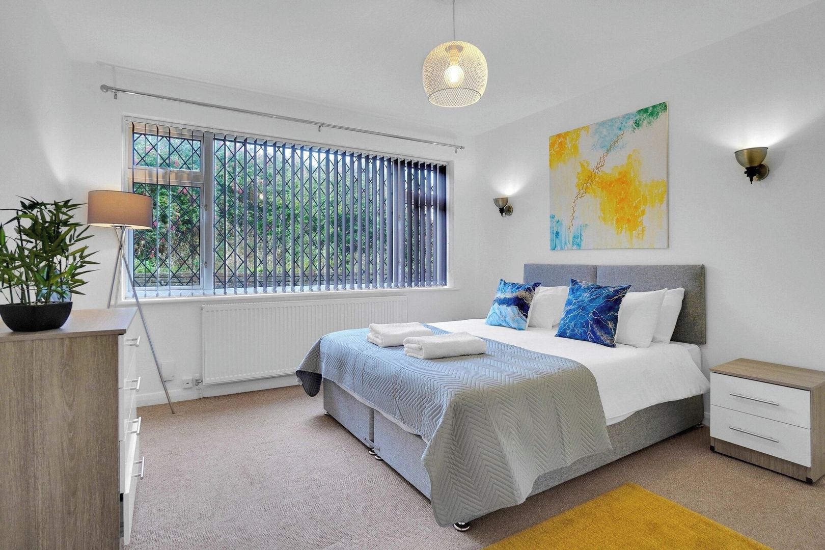 A modern bedroom with a bed, bedside tables, a window with blinds, a potted plant, and a wall-mounted light fixture.