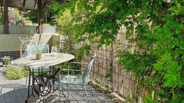 A patio with a table and chairs, surrounded by greenery and a wooden pergola.