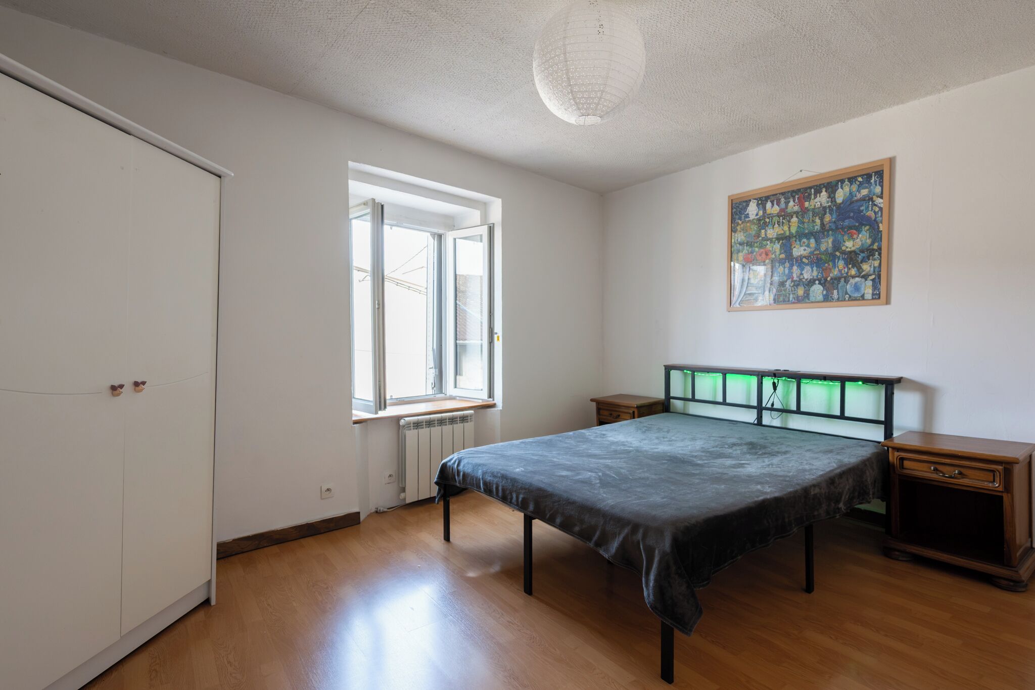 Ein Schlafzimmer mit Bett, Heizkörper, Fenster, Tür und Nachttisch.