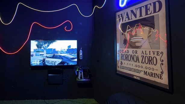 A room with a mounted TV displaying a police car, a poster with a wanted poster, and a neon light. - 70 m² Loft for 4 People* in the Jura - Patio & Arcade Room (Val Suran)