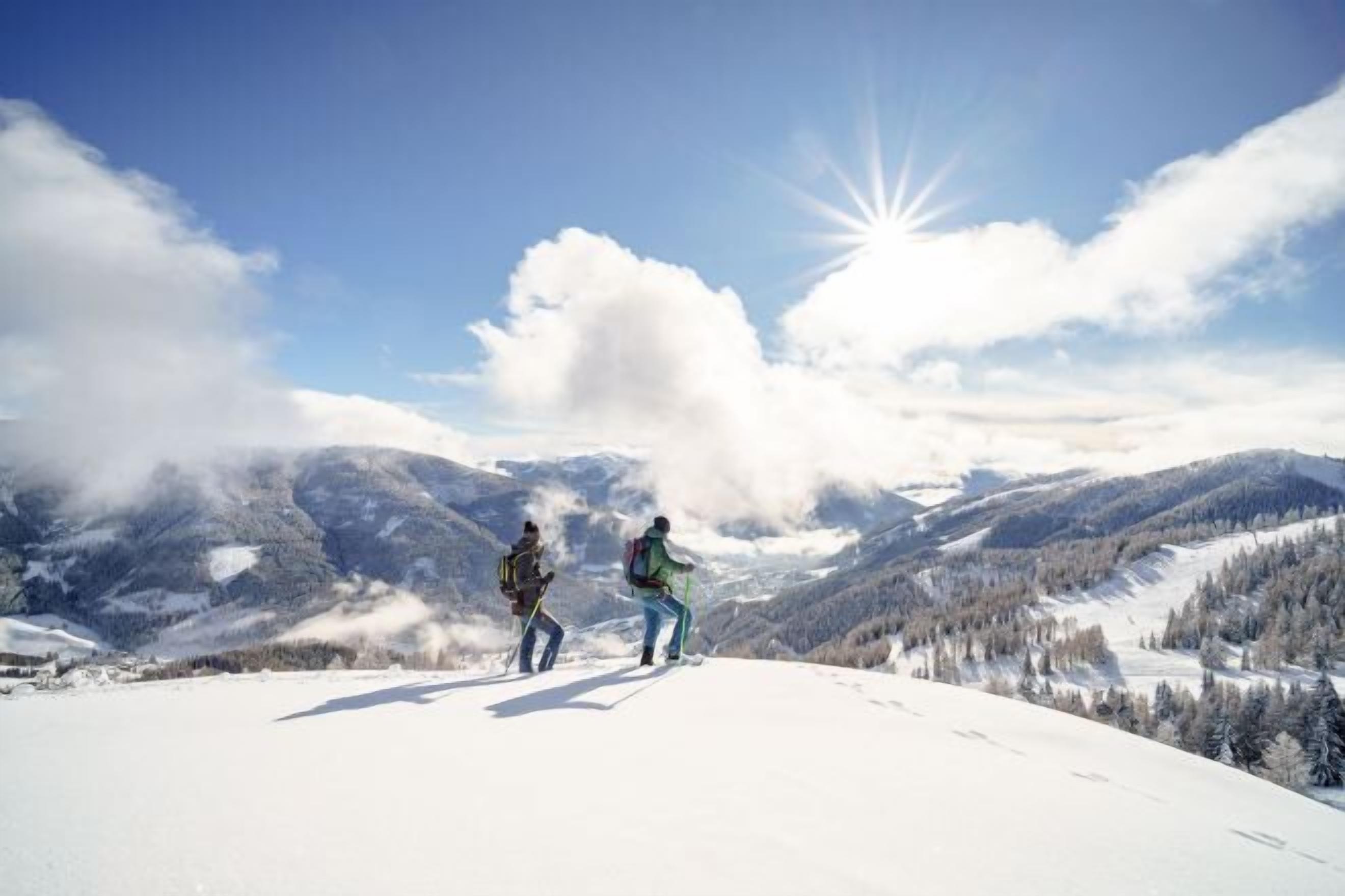 Two people hiking on a snow-covered mountain with a clear sky and sun in the background.
