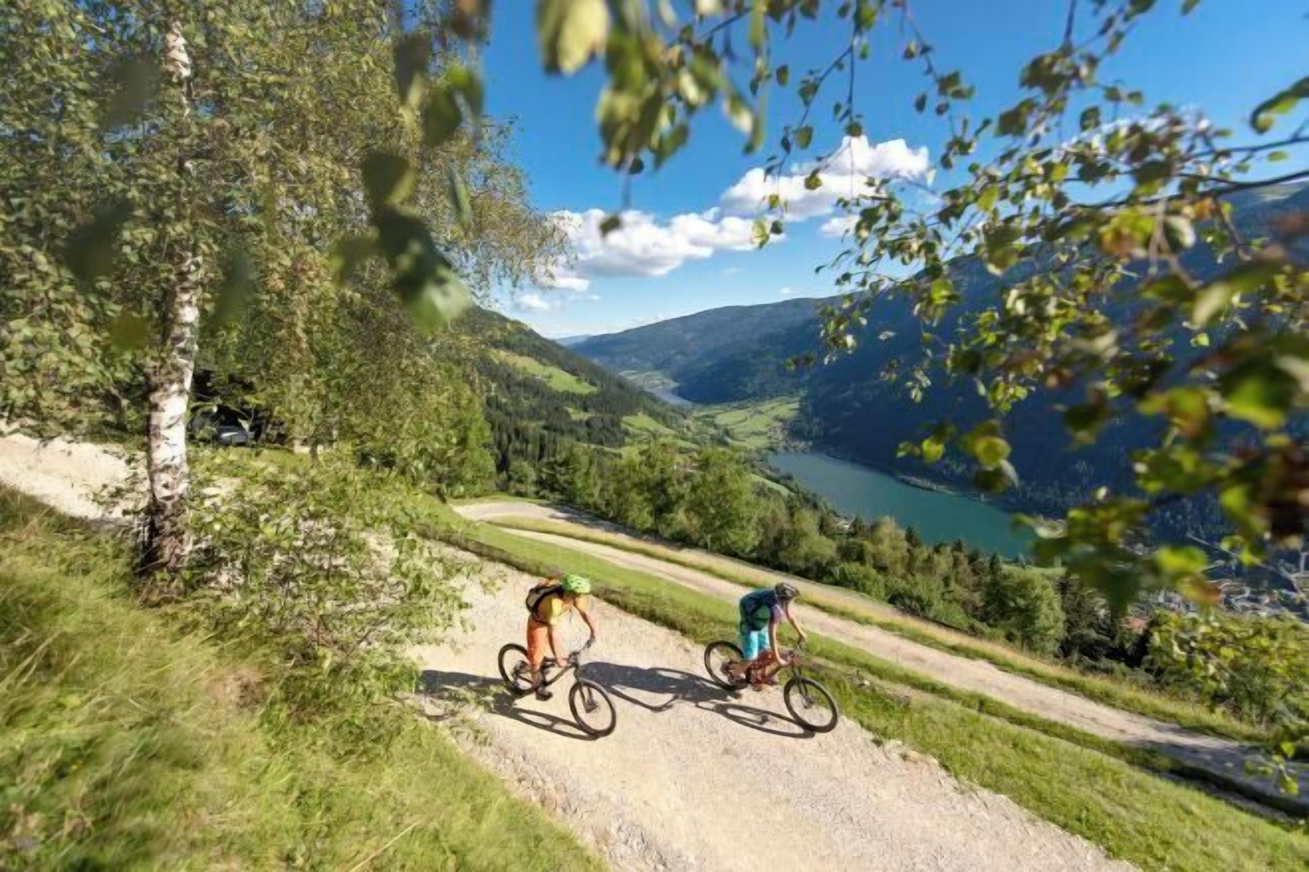 Two cyclists on a dirt path with a scenic view of a lake and mountains.