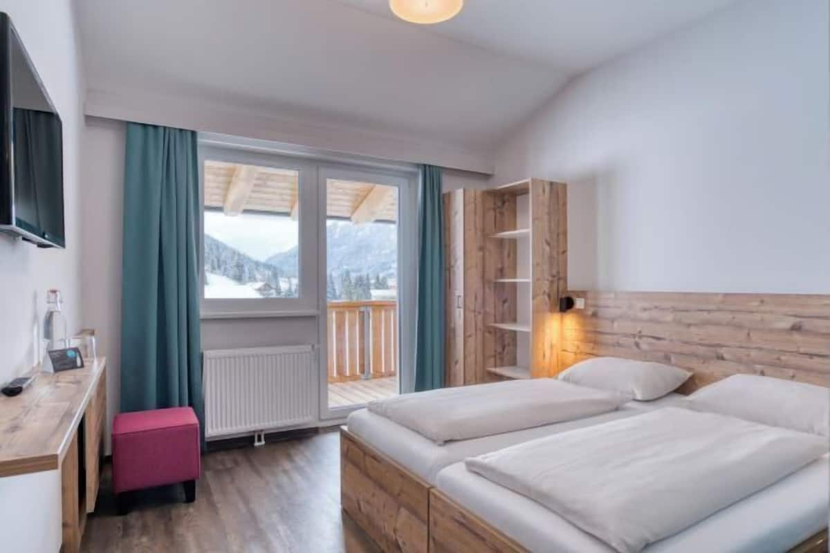 Ein Hotelzimmer mit zwei Betten, einem Holzkopfteil, einem Schreibtisch mit Stuhl, einem pinkfarbenen Hocker, einem an der Wand montierten Fernseher und Bergblick durch das Fenster.