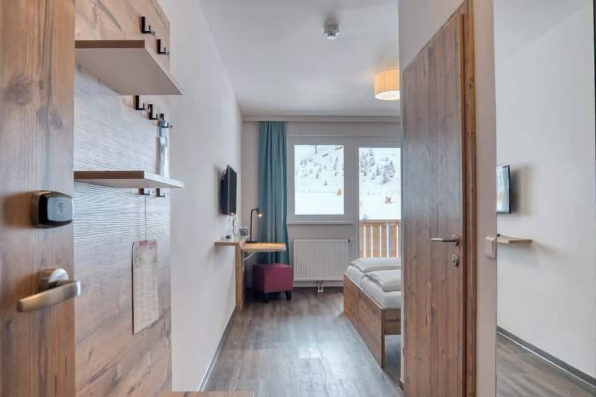 Ein modernes Hotelzimmer mit Holzboden, einem Bett, einem Schreibtisch mit Stuhl, einem Fernseher und einem Fenster mit Blick auf Bäume.