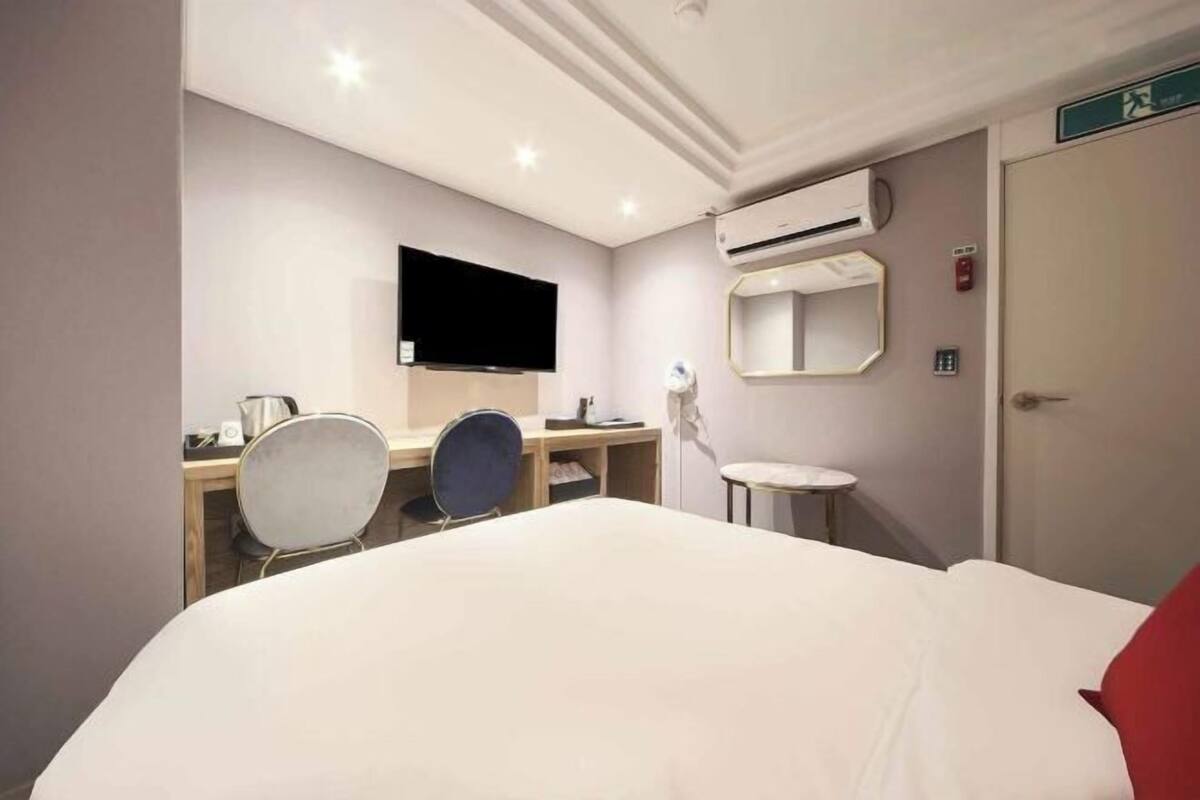 A hotel room with a bed, a desk with a chair, a television, an air conditioning unit, a mirror, and a door with an exit sign.