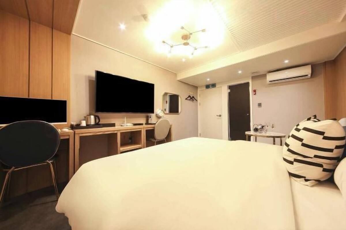 A hotel room with a bed, a desk with a flat-screen TV, a chair, a small table, and an air conditioning unit.