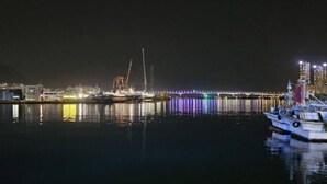 Une scène nocturne d’un port, avec un bateau amarré, une jetée illuminée et des bâtiments industriels en arrière-plan.