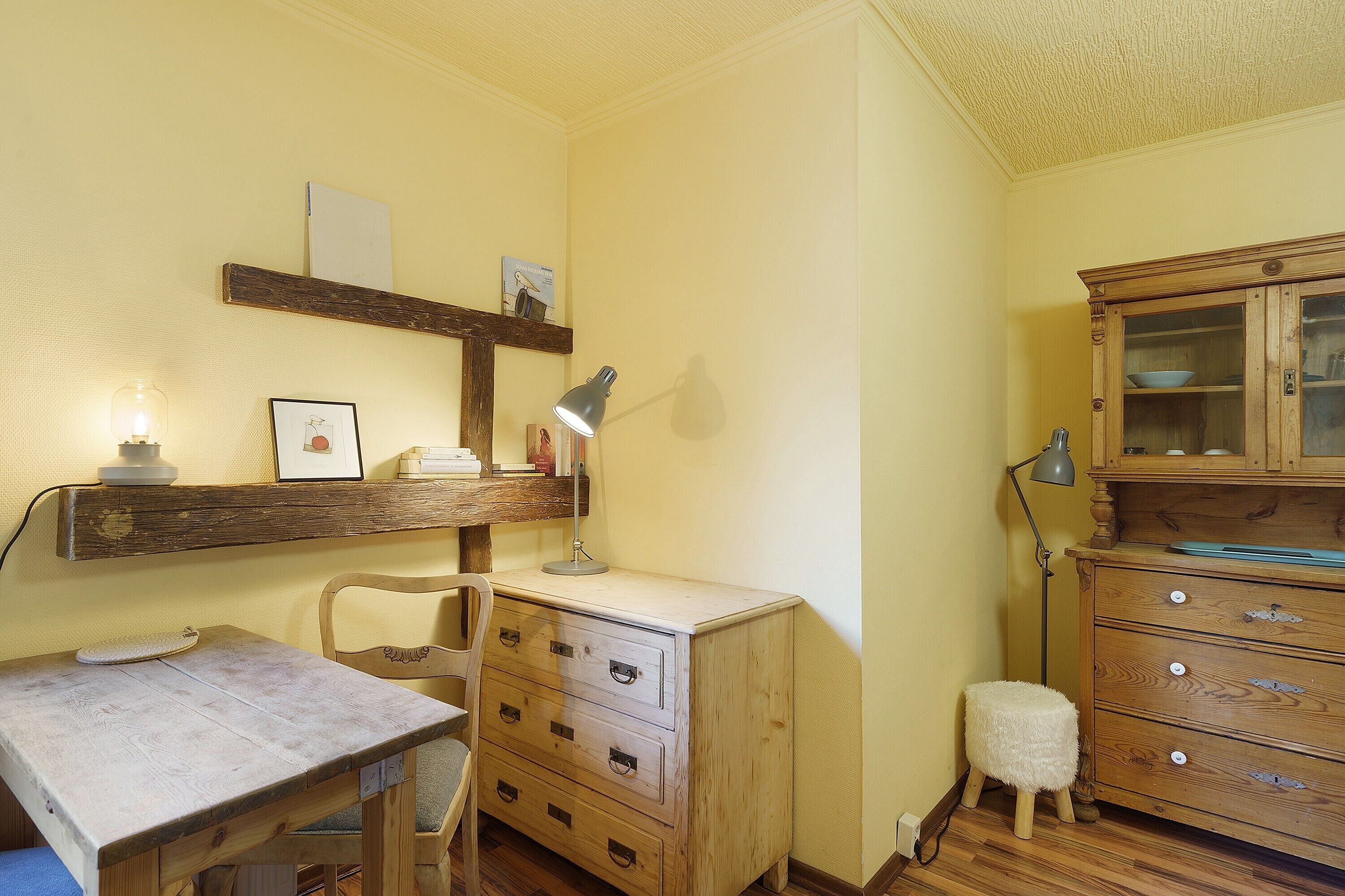 A room with a wooden desk, chair, and dresser, a small stool, and a lamp.