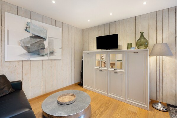 Una sala de estar moderna con televisor de pantalla plana, paneles de madera, un sofá, una mesa de centro redonda y un mueble con puertas de vidrio.