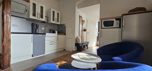 Une cuisine compacte avec des meubles blancs, un four à micro-ondes, un évier et un petit réfrigérateur. On y trouve une chaise bleue et une table ronde surmontée d’un set de table en fibres tressées.