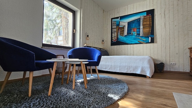 A room with a bed, two blue chairs, a small table, a painting on the wall, and a window. - Apartment 2 'Near Blue Wonder' with Shared Terrace, Garden and Wi-Fi (Dresden)