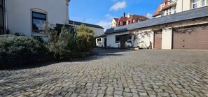 Une cour pavée, bordée de bâtiments, avec un garage et un mur blanc percé d’une fenêtre.
