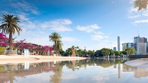 Ein Park am Wasserufer mit Palmen, einer spiegelnden Wasseroberfläche und modernen Gebäuden im Hintergrund.