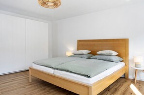 A neatly made bed with a wooden headboard, a bedside lamp, and a white wardrobe in a minimalist room.