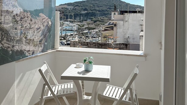 Uma varanda com mesa e cadeiras brancas, vista para uma marina e paisagem urbana ao fundo.