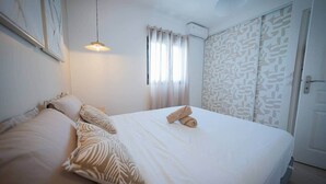 Cama bem arrumada com edredom branco e duas toalhas enroladas, luminária pendente e janela com cortinas leves.