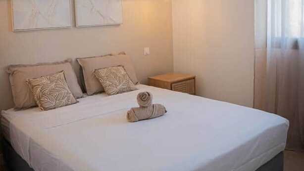 A neatly made bed with a white duvet, two pillows, and a rolled towel. There is a small bedside table with a lamp. The room has a minimalist design with plain walls and a ceiling light.