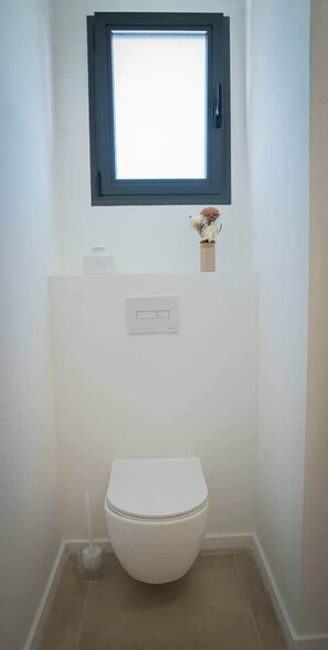 A small, clean bathroom with a white toilet, a window above it, and a small vase with dried flowers on the wall.