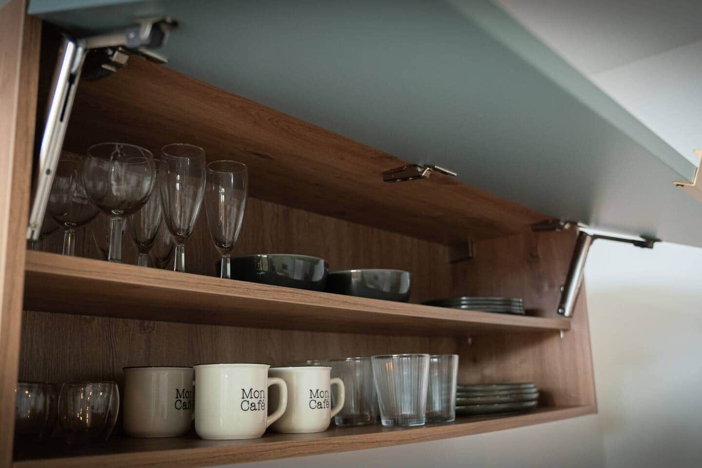 A wooden shelf with wine glasses, plates, and Mon Café mugs.