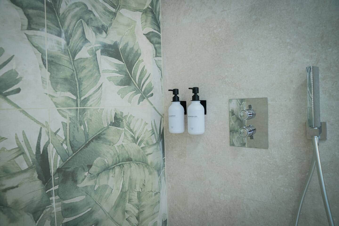 A bathroom with a shower, two bottles of shampoo and shower gel, and a wall with a leafy pattern.