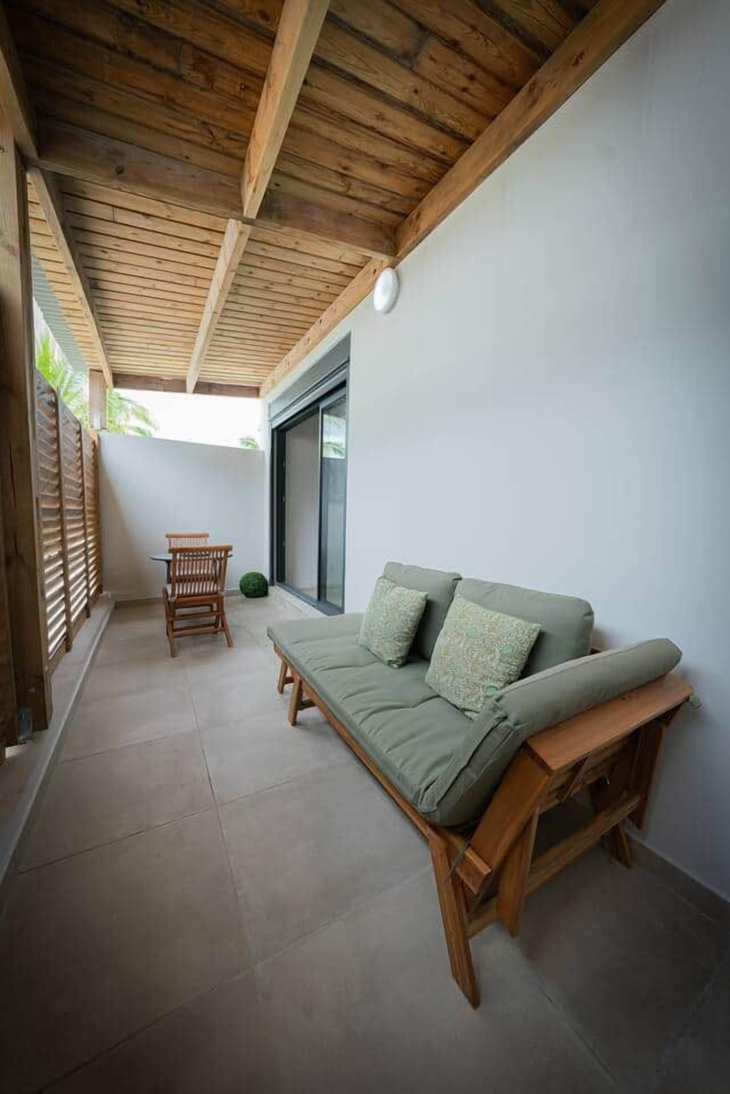 Une terrasse couverte avec un plafond en bois, un canapé vert avec des coussins, une chaise en bois et une petite table.