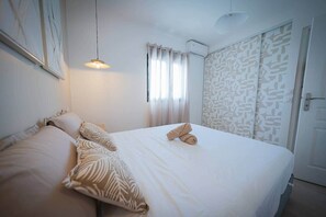 A neatly made bed with a white duvet and two rolled towels, a hanging pendant light, and a window with sheer curtains.