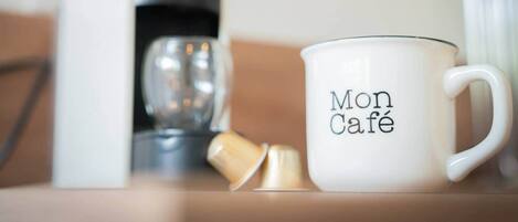 A white mug with 'Mon Café' text, a coffee maker, and a glass cup on a table.