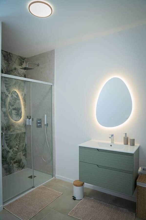 A modern bathroom with a glass shower enclosure, a green vanity, and a round mirror with integrated lighting.