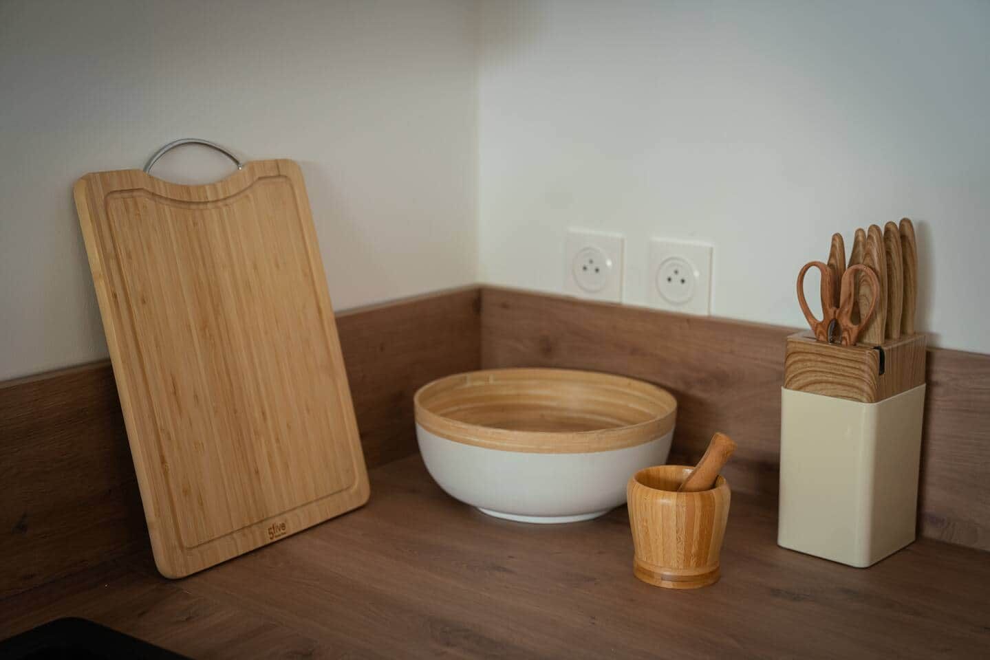 Una encimera de cocina con una tabla de cortar de madera, un tazón blanco, una cuchara de madera en un portalápices y un bloque de cuchillos.