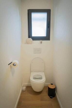 A small, clean bathroom with a toilet, a window, a toilet paper holder, and a trash bin.