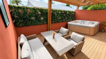 Un patio al aire libre con mesa de madera, sofá de mimbre, bañera de hidromasaje y un muro cubierto de seto verde.