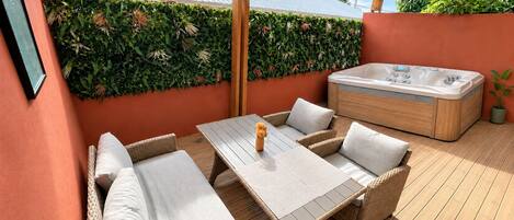 Een terras buiten met een houten tafel en een rieten bank, een jacuzzi en een groene haagwand.