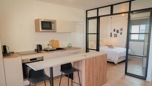 A modern kitchen with a microwave, sink, and bar stools. There is a bed with a white bedspread in the adjacent room. - Cosy T2 - La Verrière (La Possession)