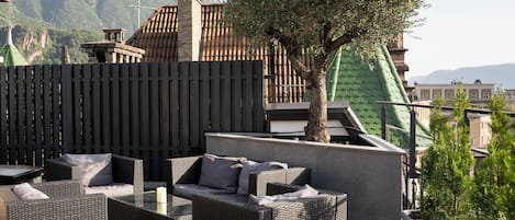 An outdoor seating area with wicker furniture and a tree, set against a backdrop of mountains and buildings.