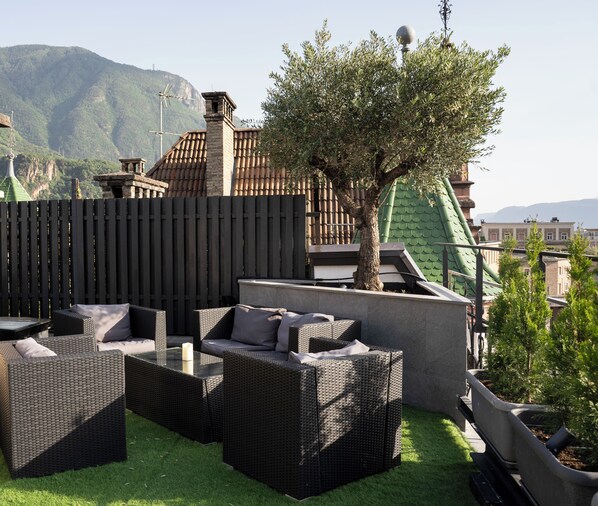An outdoor seating area with wicker furniture and a tree, set against a backdrop of mountains and buildings. - Talvera Dream (Bolzano)