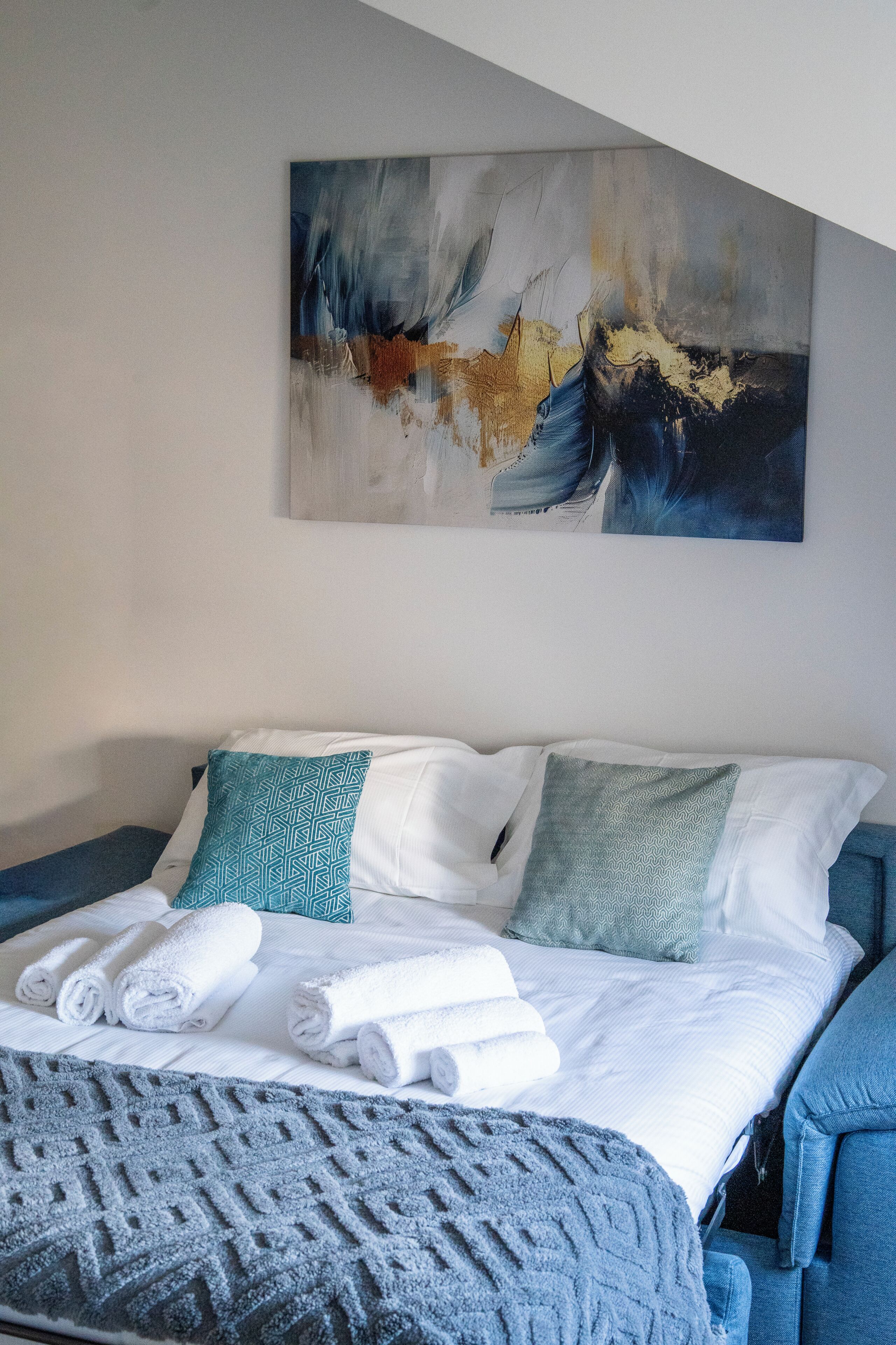 A hotel room with a bed, pillows, a blanket, and a painting on the wall.