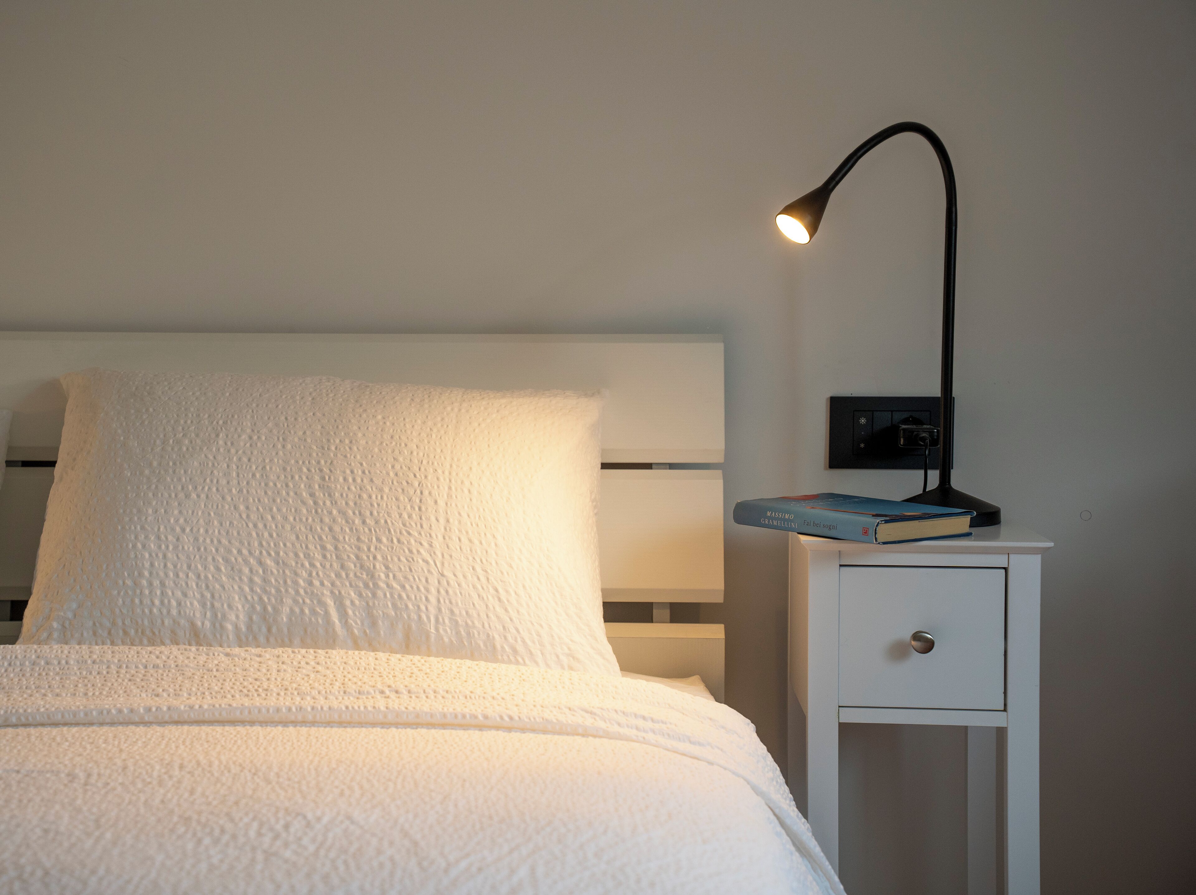 Un lit bien fait, avec un oreiller blanc et une couverture blanche, une table de chevet avec une lampe et un livre intitulé « Massimo Gravellini ».
