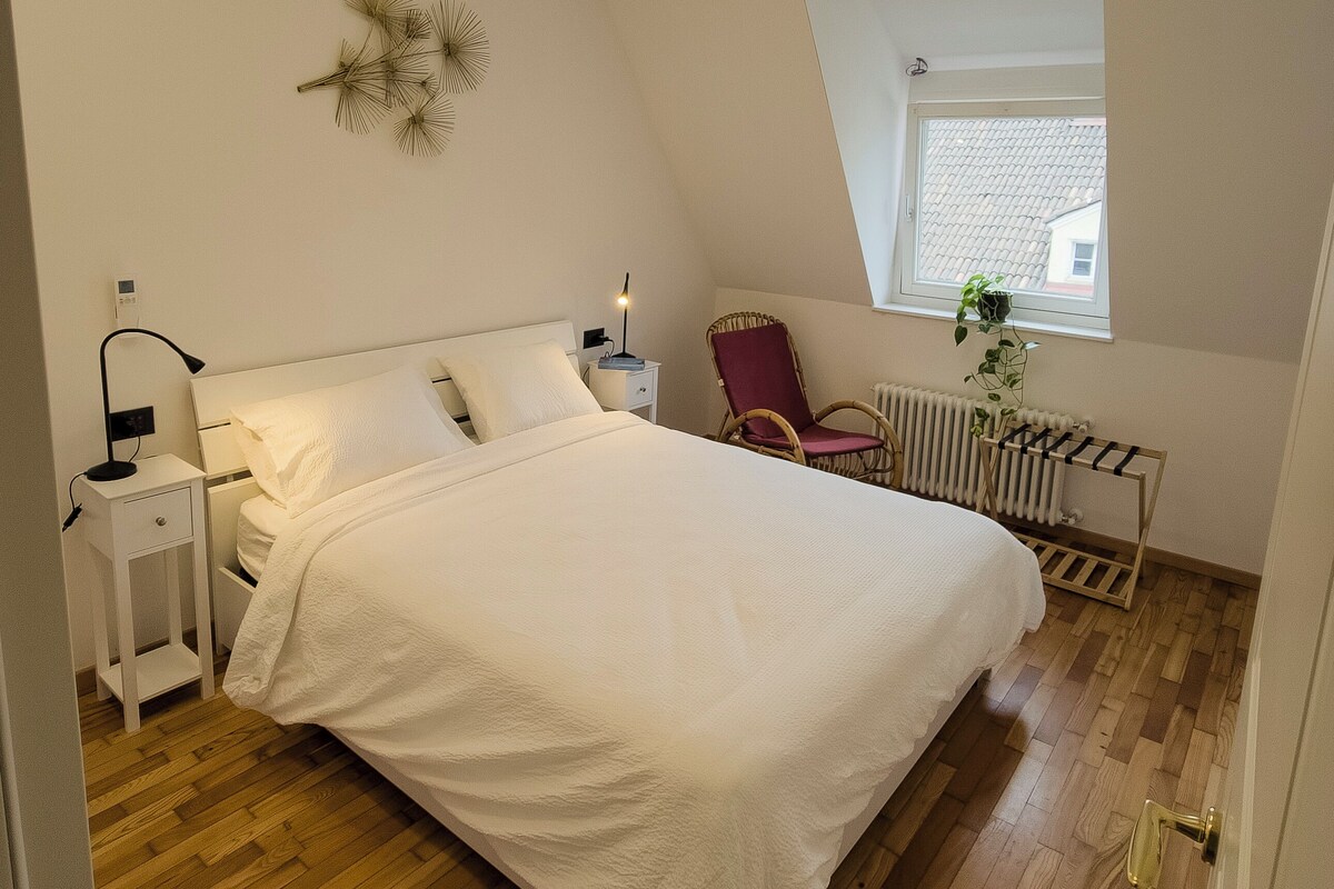 A bedroom with a large bed, a nightstand, a chair, a radiator, and a window with a view of the outside.