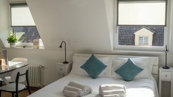 A bedroom with a large bed, two bedside tables, a desk, and a chair. There are two windows with a view of a house.