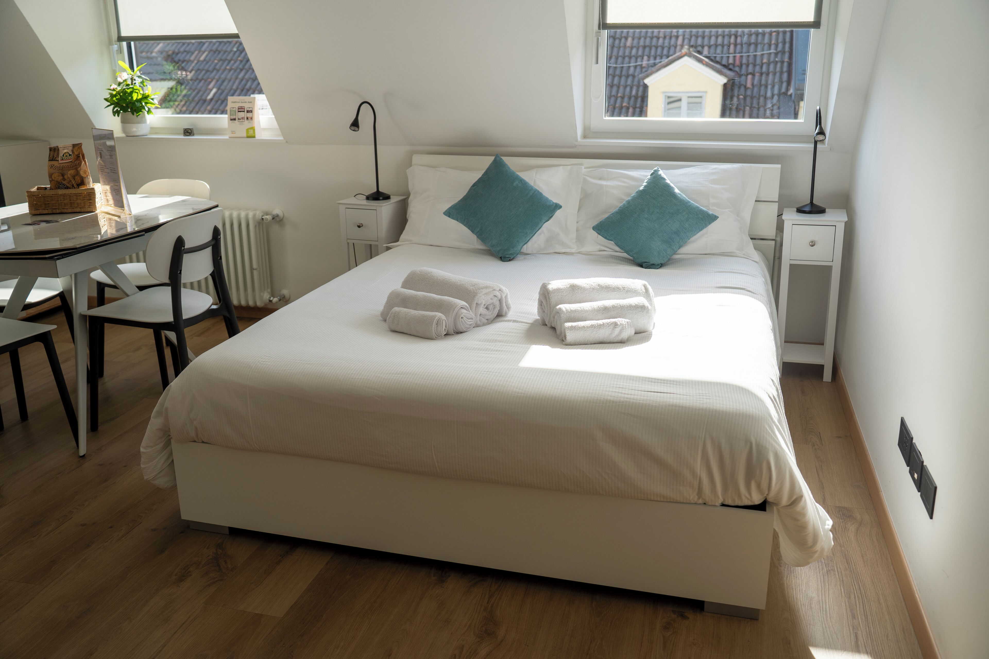 A hotel room with a large bed, two pillows, a desk with a chair, a radiator, and a window with a view of a house.