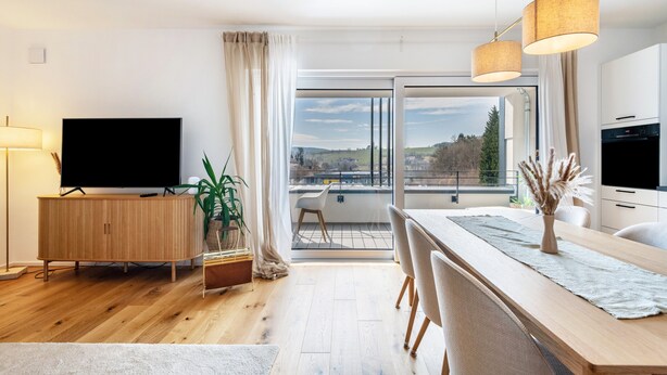 A modern living room with a flat-screen TV, wooden cabinet, dining table, chairs, and a view through large windows. - Apartment 'Bei Stefanie' with Mountain View, Balcony and Wi-Fi (Bad Kötzting)
