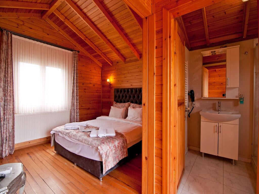 Un dormitorio de cabaña de madera con una cama, una mesita de noche, una ventana con cortinas y un baño con lavamanos y armarios.
