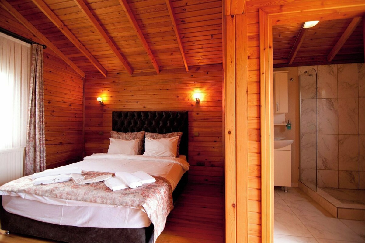 A wooden cabin bedroom with a bed, headboard, bedside lamps, and a bathroom with a shower and sink.