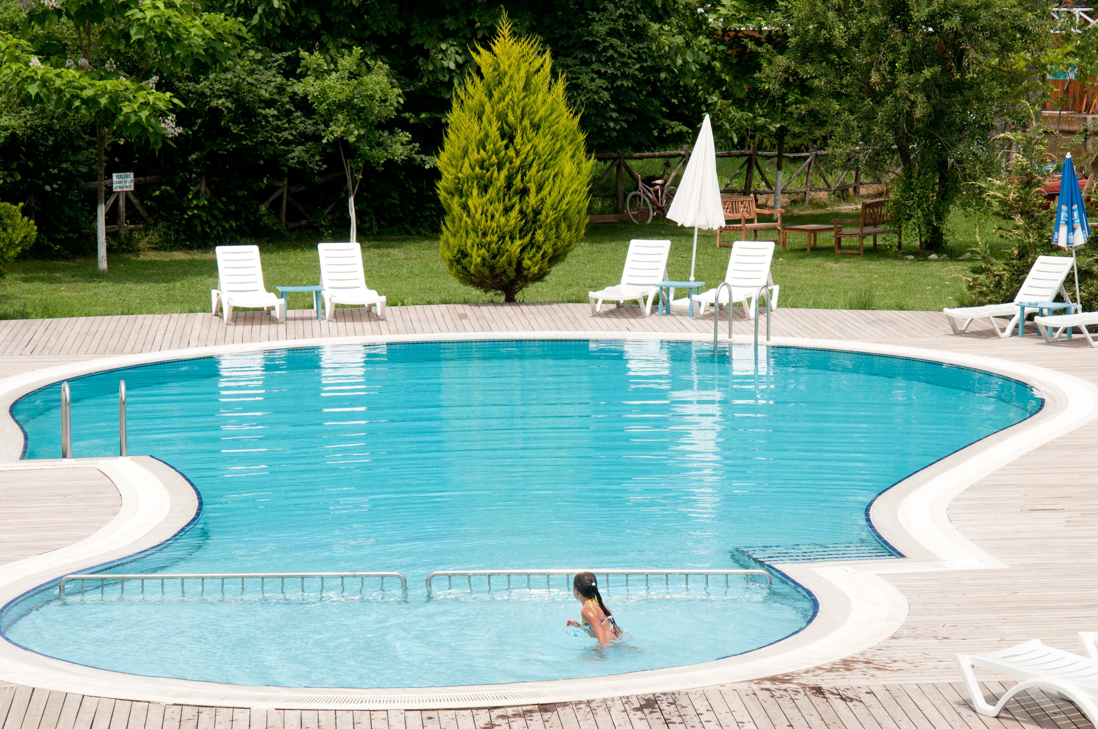 A swimming pool with a wooden deck, surrounded by white lounge chairs and a small table with a white umbrella.