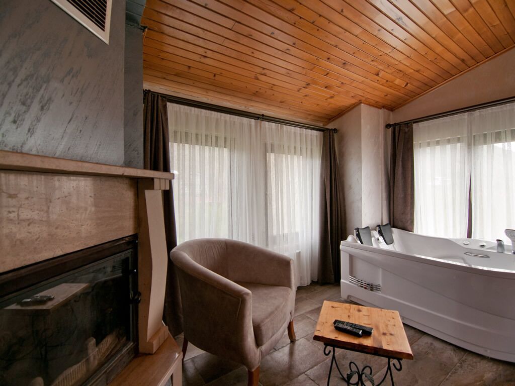 A room with a wooden ceiling, a fireplace, a large window with curtains, a bathtub, and a remote control on a table.