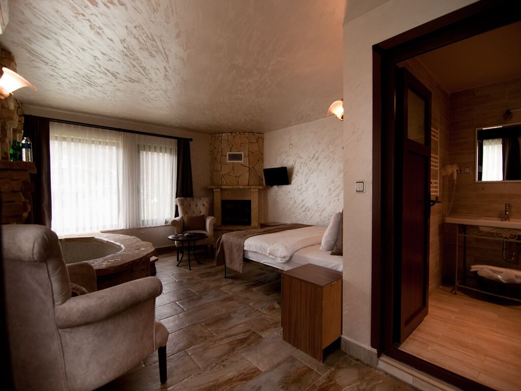 A room with a bed, a chair, a small table, a fireplace, and a bathroom with a sink and mirror.