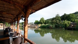 Un restaurante al aire libre con mesas y sillas de madera, con vista a un río tranquilo, vegetación y un puente al fondo.